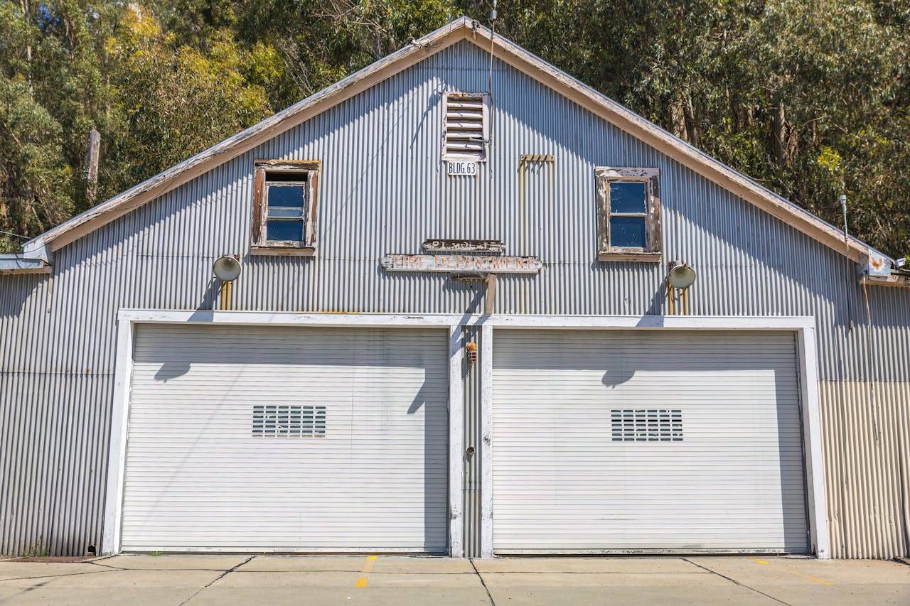gallery-1 A sunlit vintage metal garage with double doors and rustic charm amidst trees.