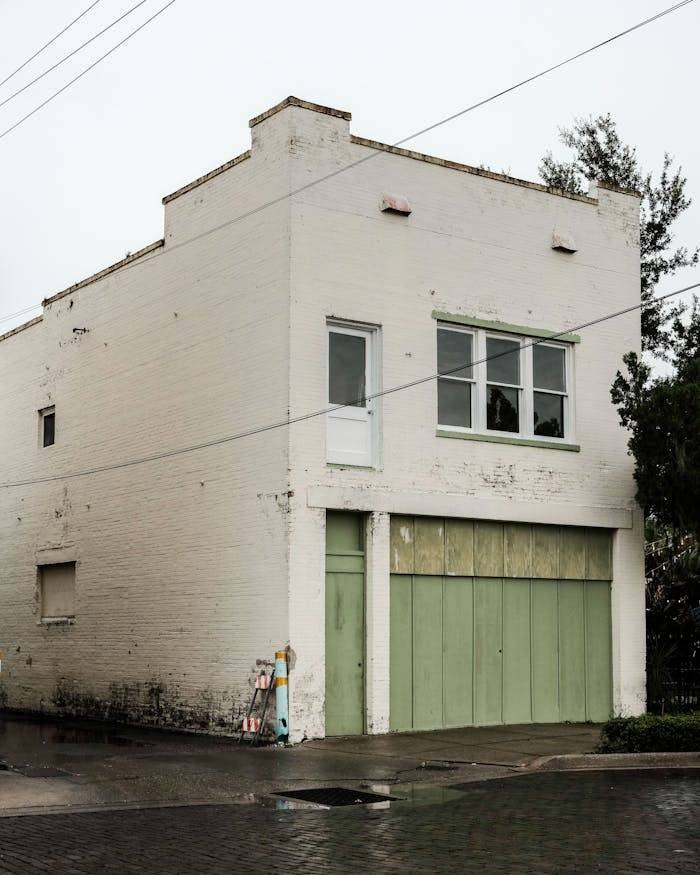 gallery-6 Aged white brick building with green garage doors on a rainy street corner, evokes a vintage urban feel.