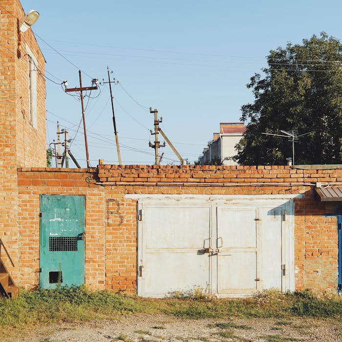 Services-02 A rustic brick wall featuring a green door and white garage door under clear skies, casting distinct shadows.