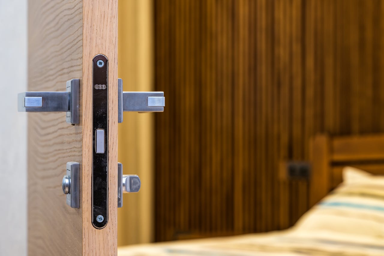Opened doorway with a lock and simple metal handle leading into a light room with a wooden bed